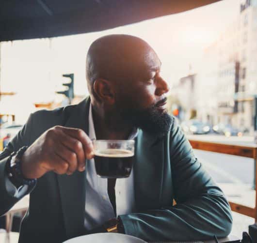 A square shot of a handsome black man drinking coffee in a street cafe on a sunny morning; the portrait of a dapper African American guy looking aside while having a coffee in an outdoor restaurant
